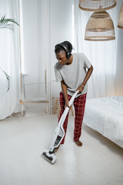 A close-up view of a red portable vacuum cleaner with a black power cord, placed on a beige textured area rug in a domestic setting. The vacuum is positioned near a wooden cabinet with a natural finis