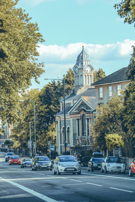 A street scene in Barnes displaying a mix of residential and commercial buildings with mature trees lining the sidewalk. In the background, a historic clock tower featuring a tall spire and ornate arc